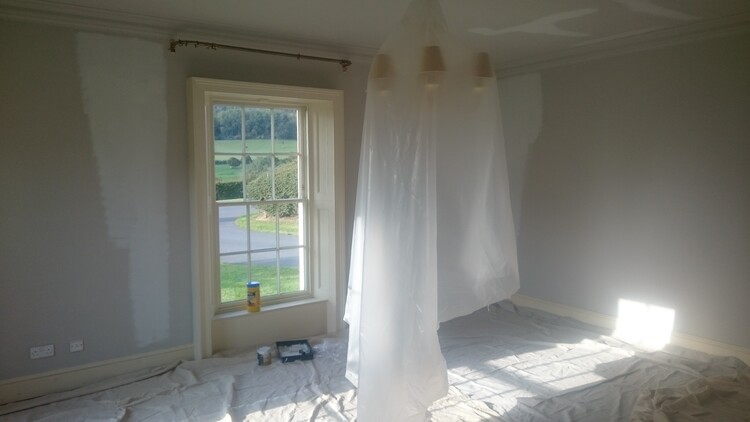 A room with tall sash windows is painted, with dust sheets protecting the chandelier and wooden floor.